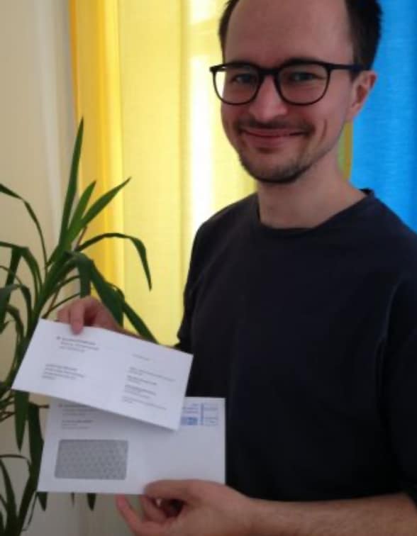 Mann mit Brille hält Briefe in der Hand, Pflanze und Vorhang im Hintergrund.