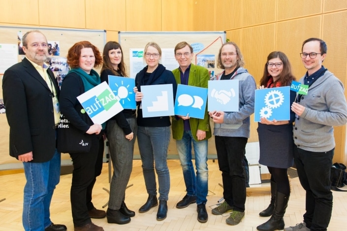 Gruppe von Personen hält Schilder mit Symbolen und dem Logo aufZAQ. Feierliche Veranstaltung.