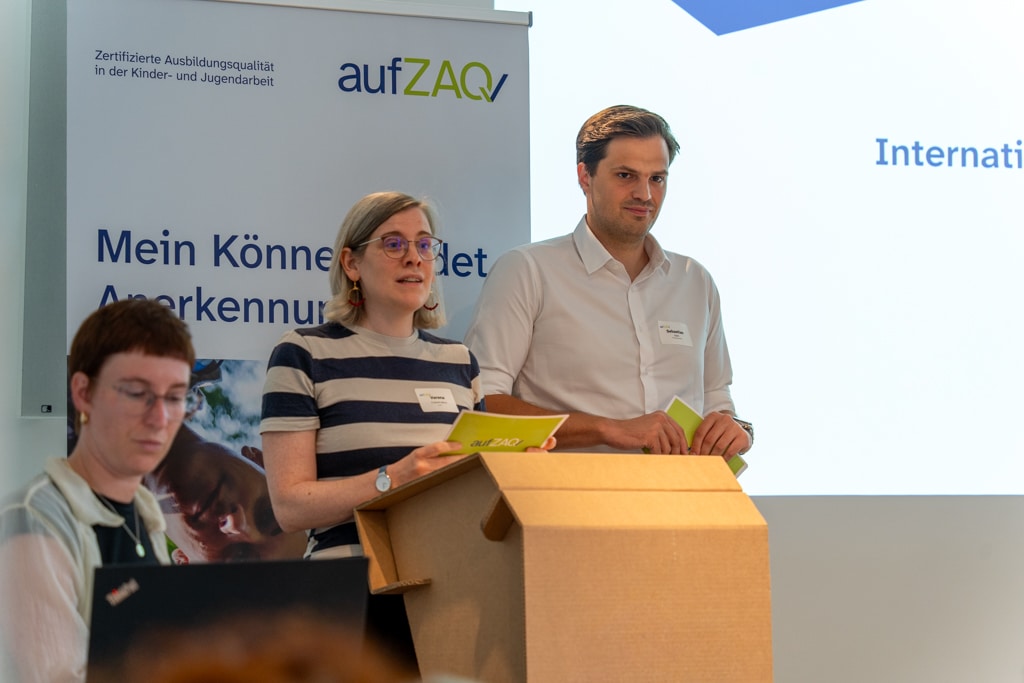 Zwei Personen präsentieren auf dem aufZAQ Impuls.Brunch vor Publikum. Eine Person tippt am Laptop.